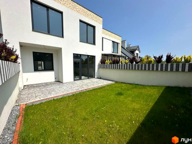 Фасад білого будинку з великими вікнами та зеленою галявиною перед ним. Територія огороджена парканом з рослинами.