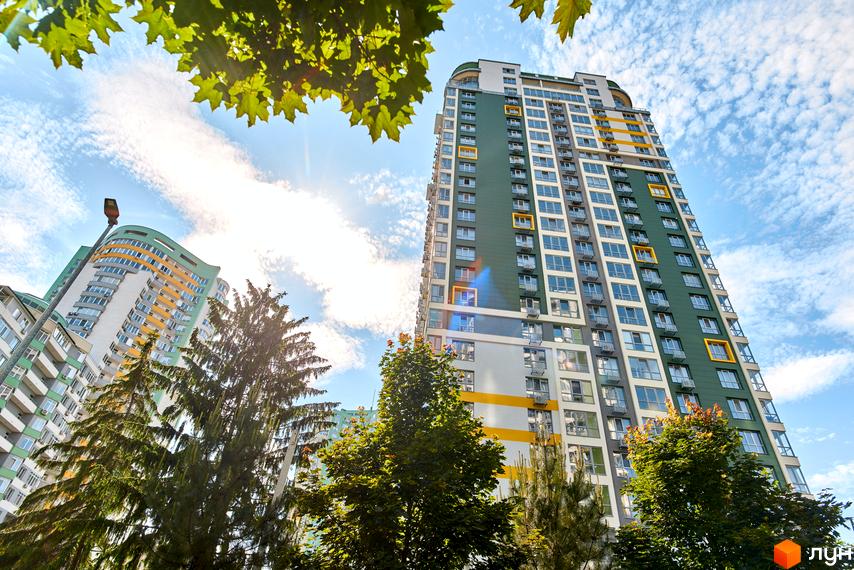 Житловий комплекс Паркове місто: високі житлові будинки з фасадами темно-зеленого, білого та жовтого кольорів. Навколо будівель багато зелених дерев під блакитним небом.