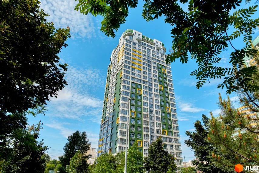 Житловий комплекс Паркове місто: високий будинок з біло-зеленим фасадом та жовтими акцентами на вікнах, оточений густою зеленню дерев під блакитним небом.