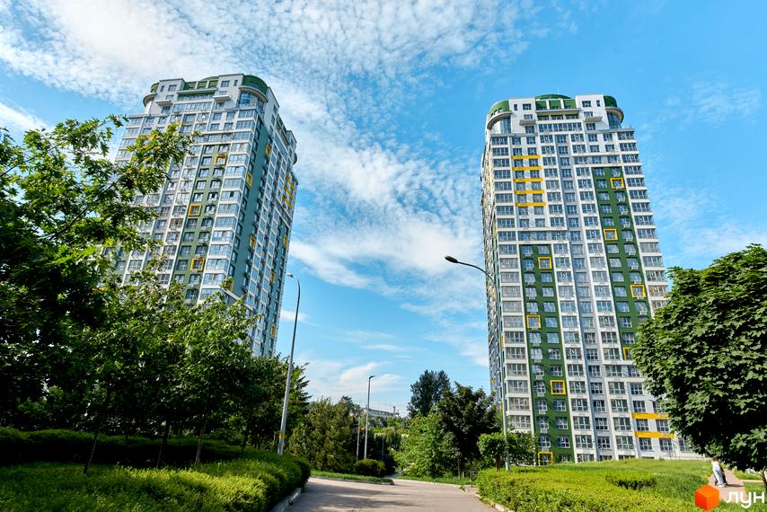 Житловий комплекс Паркове місто: дві високі житлові будівлі з біло-зелено-жовтими фасадами та численними вікнами, оточені зеленими насадженнями та доріжкою.