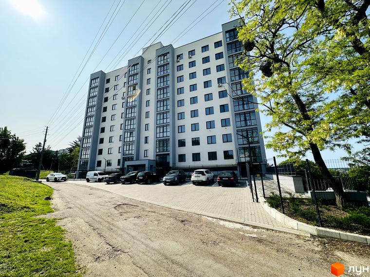Житловий комплекс Соборний: багатоповерховий будинок світлого кольору з темними акцентами. Прибудинкова територія з паркінгом, зеленими насадженнями та під'їзною дорогою.