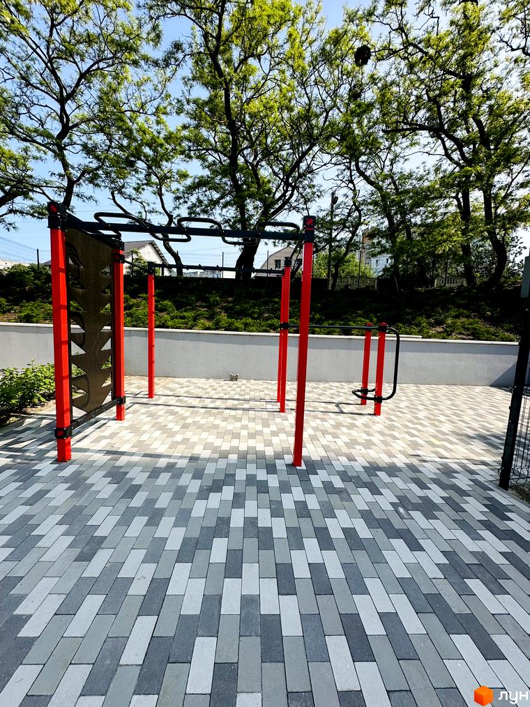 Житловий комплекс Соборний: на прибудинковій території облаштовано спортивний майданчик з червоними тренажерами на бруківці. На фоні видно дерева та житлові будівлі.