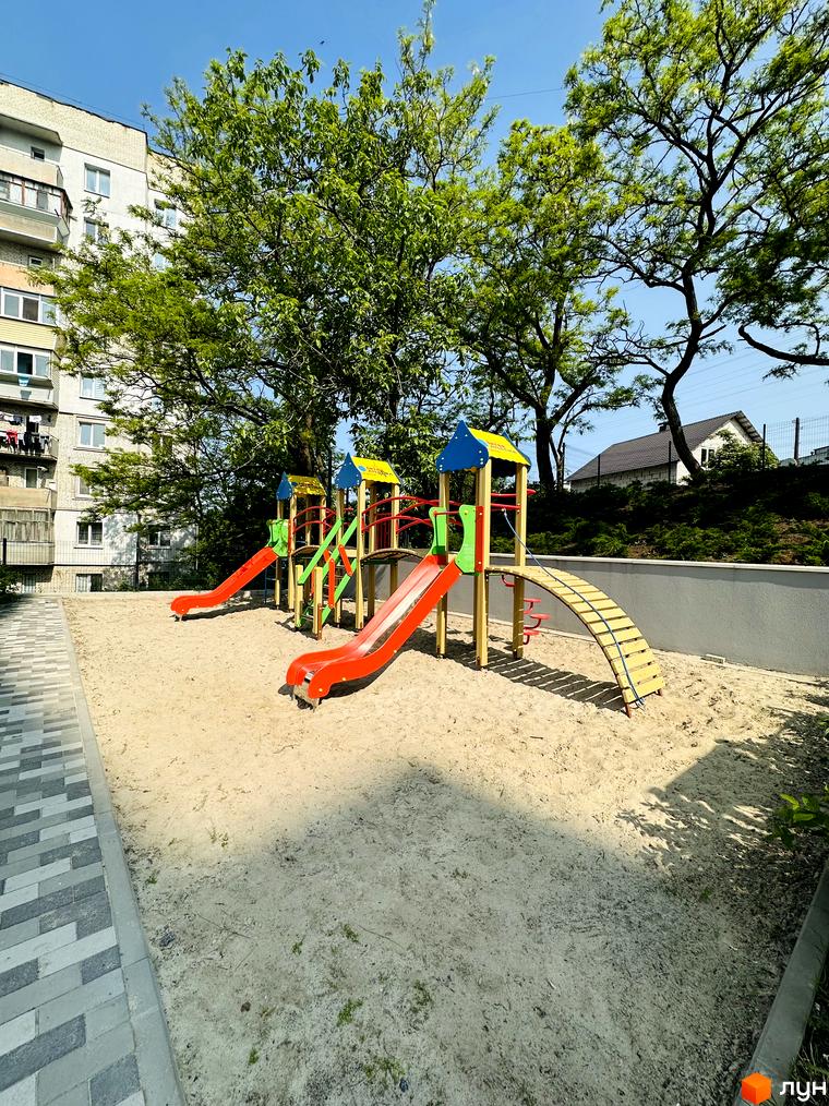 Житловий комплекс Соборний: багатоповерховий будинок світлого кольору з балконами. На прибудинковій території піщаний дитячий майданчик з гірками та деревами.