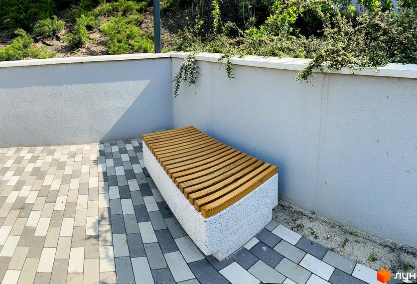 Прибудинкова територія Житлового комплексу Соборний: лавка з дерев'яним сидінням, мощена доріжка з плитки та зелені кущі вздовж стіни.