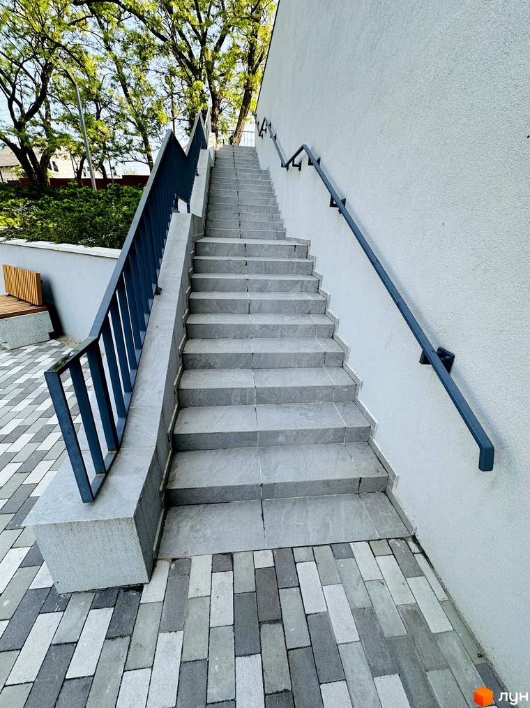 Вхідні сходи з сірої плитки та темними поручнями біля світлого фасаду Житлового комплексу Соборний. Територія має бруківку та зелені насадження.