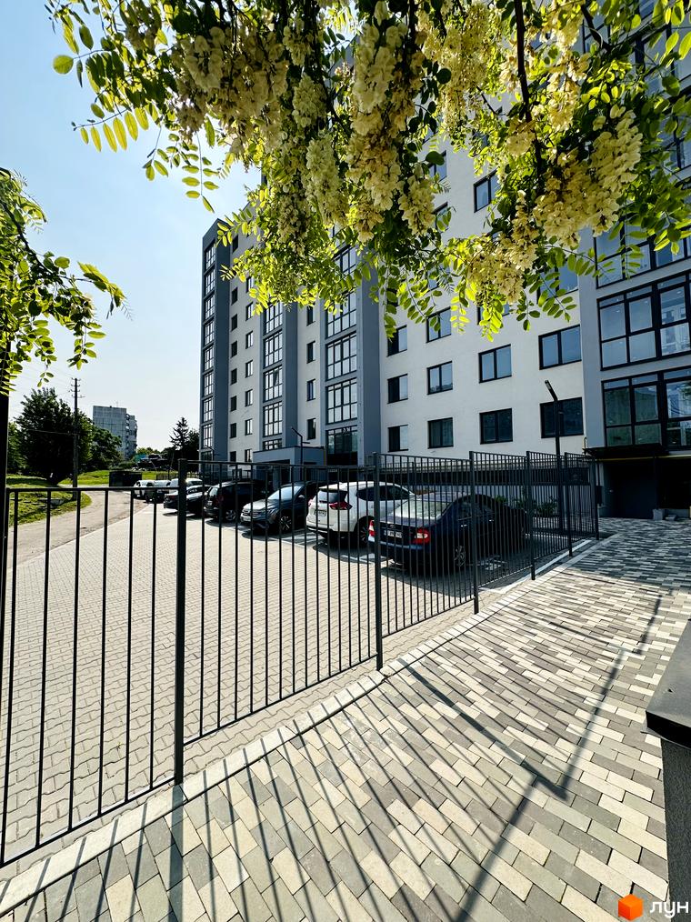 Багатоповерховий будинок Житлового комплексу Соборний з білим фасадом та темними вікнами. Перед будинком огороджена парковка з автомобілями та вимощена тротуарна плитка.