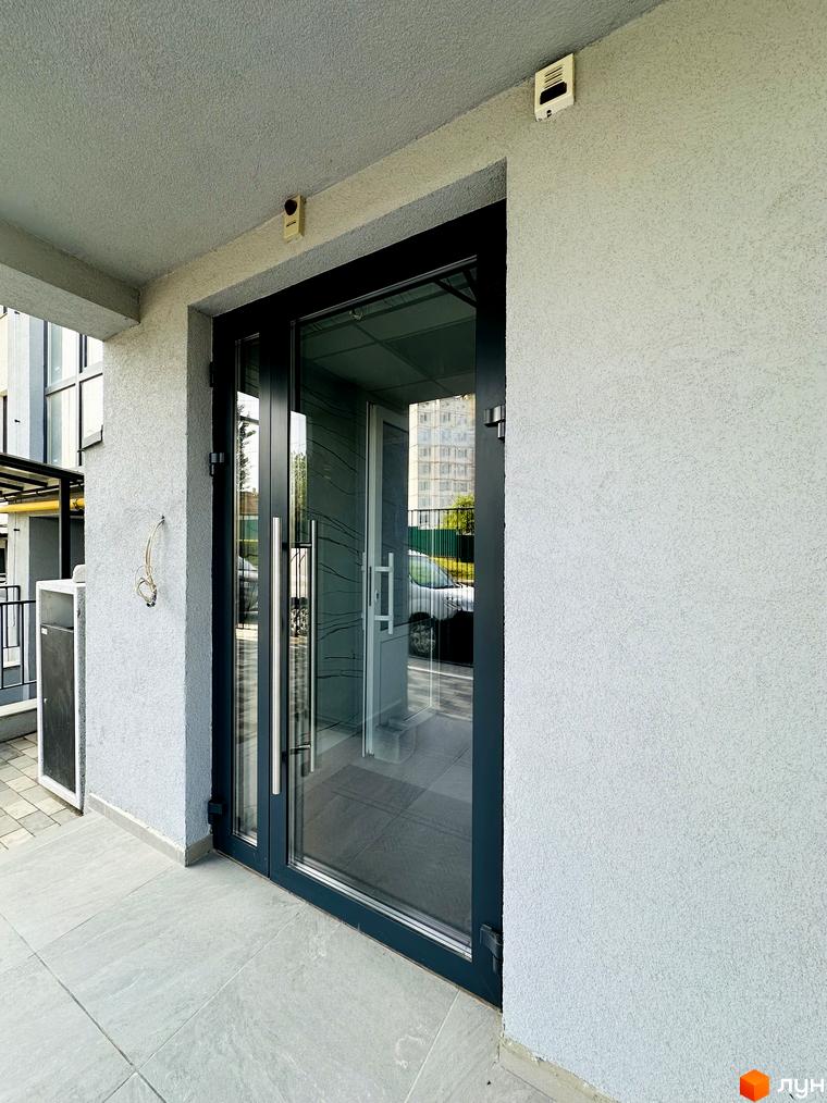 Вхід до Житлового комплексу Соборний з світло-сірим фасадом та великими скляними дверима. Перед входом плитка на підлозі та вид на прибудинкову територію.