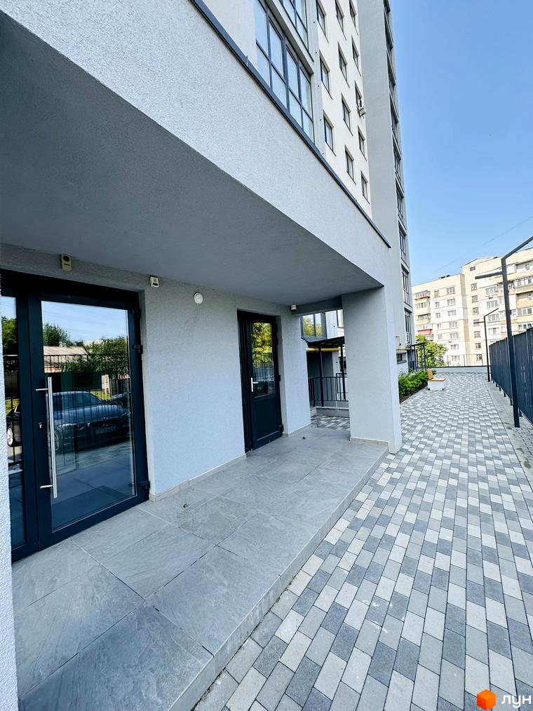 Сірий багатоповерховий будинок Житловий комплекс Соборний з великими вікнами та темними дверима на першому поверсі. Прибудинкова територія вимощена сірою плиткою.