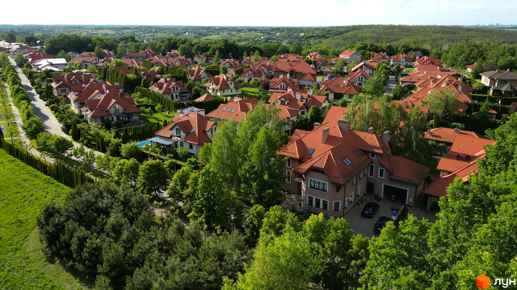 Зовнішній вигляд котеджного містечка КМ Рославичі з червоними дахами та зеленими насадженнями. Прибудинкова територія оформлена деревами та газонами.