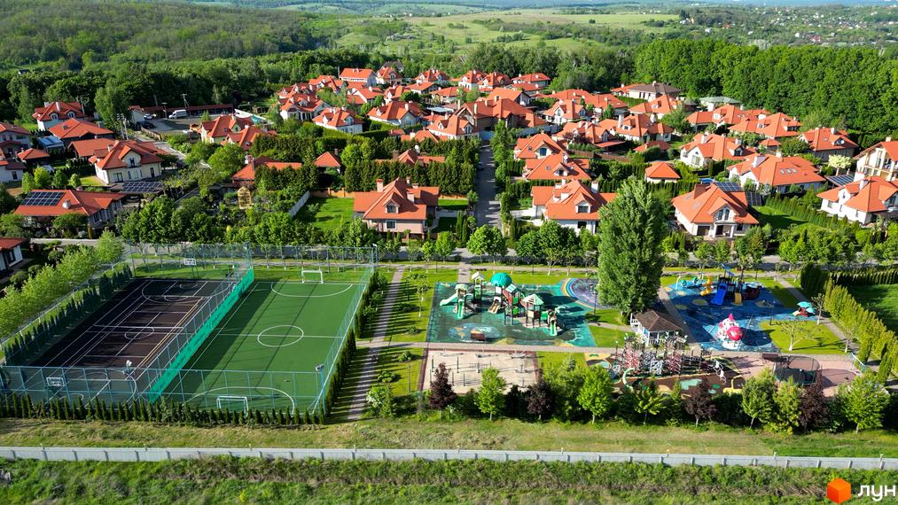 Вигляд житлового комплексу КМ Рославичі з котеджами з червоною покрівлею та зеленою прибудинковою територією з дитячими майданчиками.