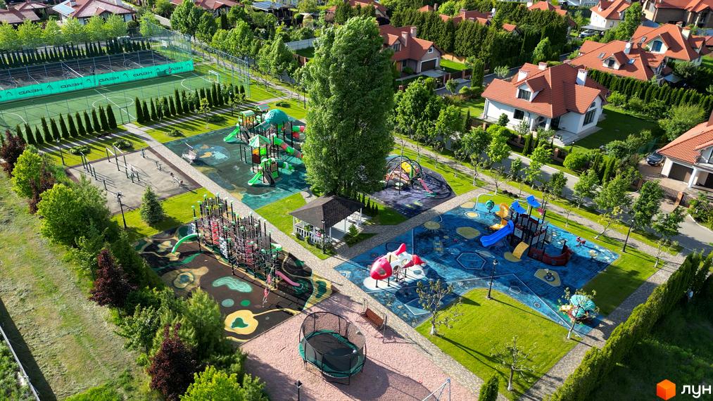 Вигляд з висоти на Котеджне містечко Рославичі: дитячі майданчики, спортивне поле та зелені насадження. Будинки з червоними дахами серед дерев.