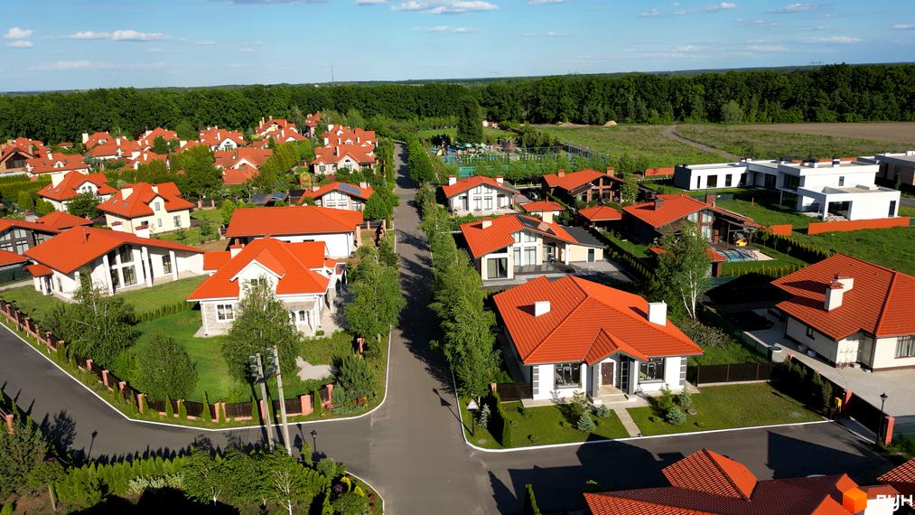 Фото Котеджного містечка Рославичі з виглядом на вулицю з будинками з червоними дахами. Навколо будинків зелені газони та дерева.