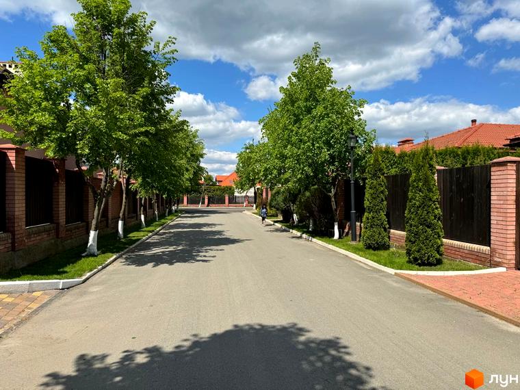 Вулиця в Котеджному містечку Рославичі з деревами вздовж дороги. Охайні будинки з червоними дахами та зелені насадження.