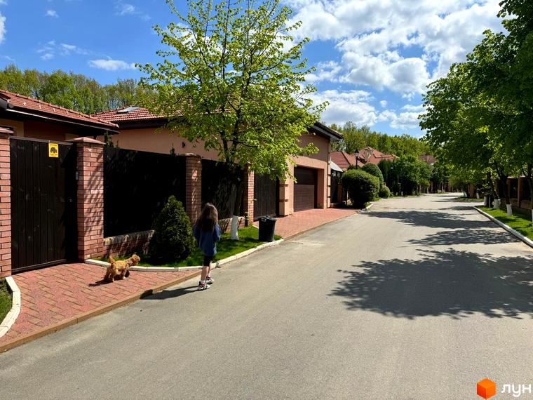 Вигляд на Котеджне містечко Рославичі з вулиці. Будинки з червоними дахами, зелені дерева вздовж дороги, дитина з собакою на тротуарі.