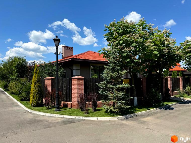 Зображення будинку в Котеджному містечку Рославичі з червоним дахом та зеленим подвір'ям. Навколо будинку дерева та декоративні рослини.