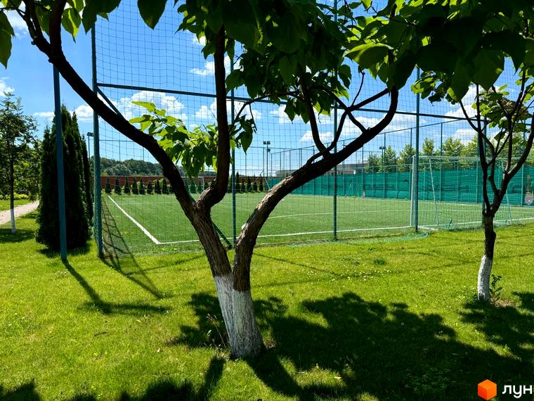 Зображення футбольного поля в Котеджному містечку Рославичі. На передньому плані дерева з зеленим листям, газон та огорожа.
