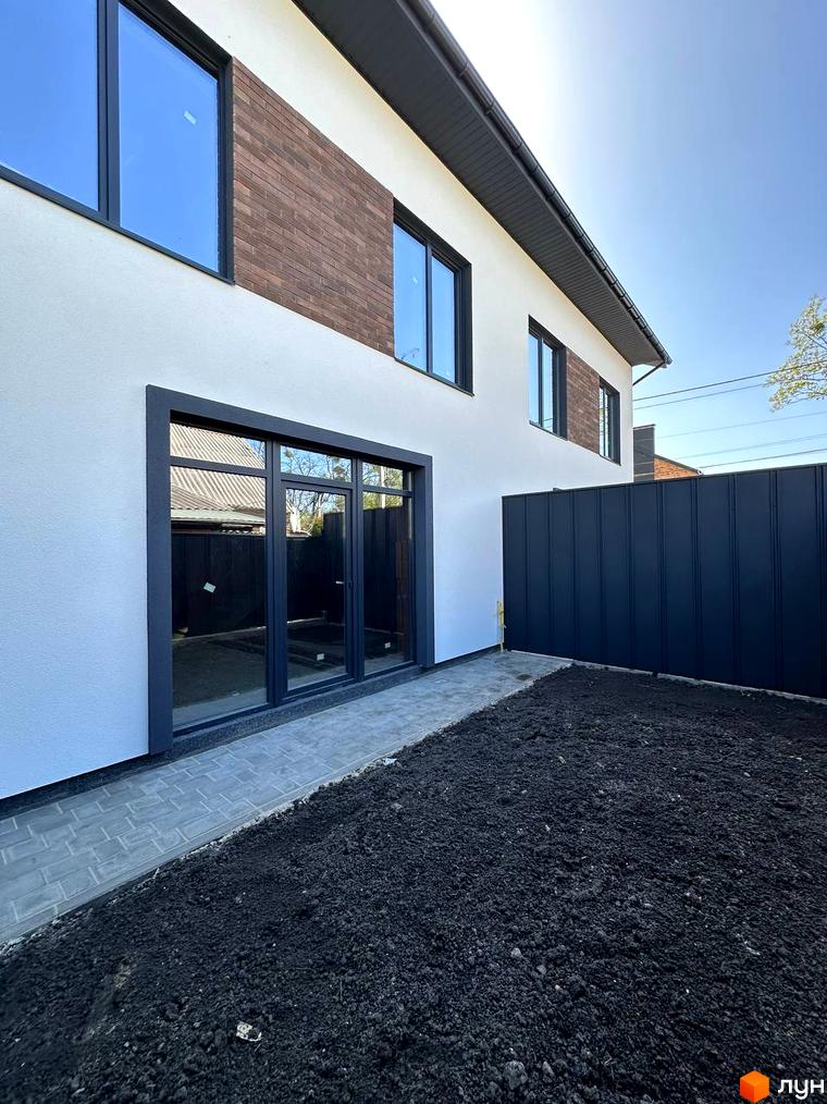 Таунхауси „В13“: білий фасад з цегляними вставками, великі вікна та вхідна група, огороджена чорним парканом. Поруч викладена доріжка з плитки та ділянка з чорноземом.