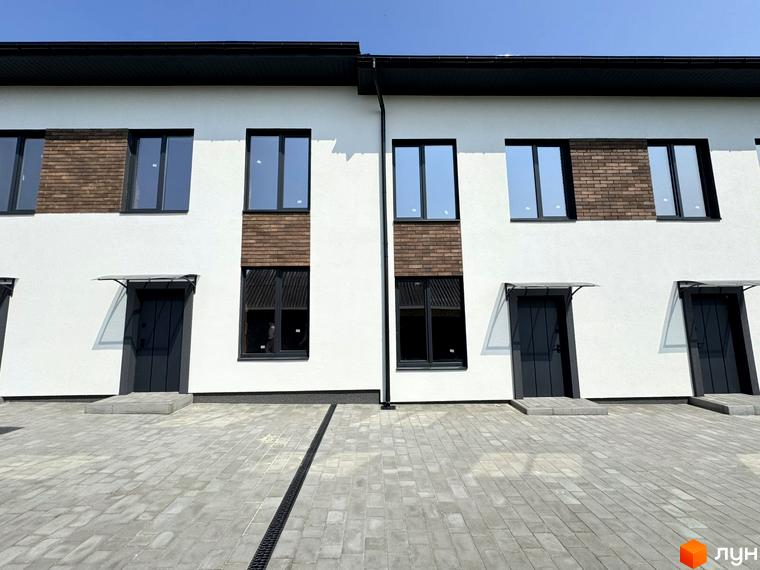 Фасад таунхаусів „В13“ з білими стінами, темними вікнами та дверима, оздоблені декоративною цеглою. Перед будинками викладена бруківка.