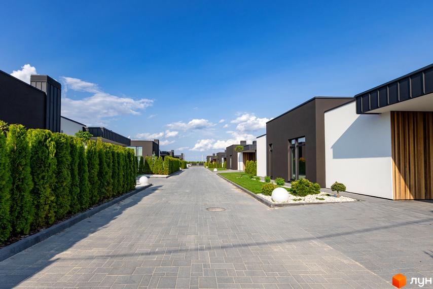 Дорога в Містечко котеджів Terra Solis з сучасними будинками з темними фасадами, білими стінами та доглянутою прибудинковою територією з рослинами та ліхтарями.