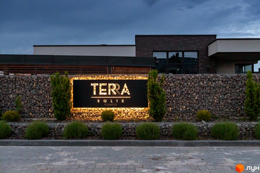 Котеджне містечко Terra Solis: фасади будівель та освітлена вивіска на фоні кам'яної огорожі та зелених насаджень.