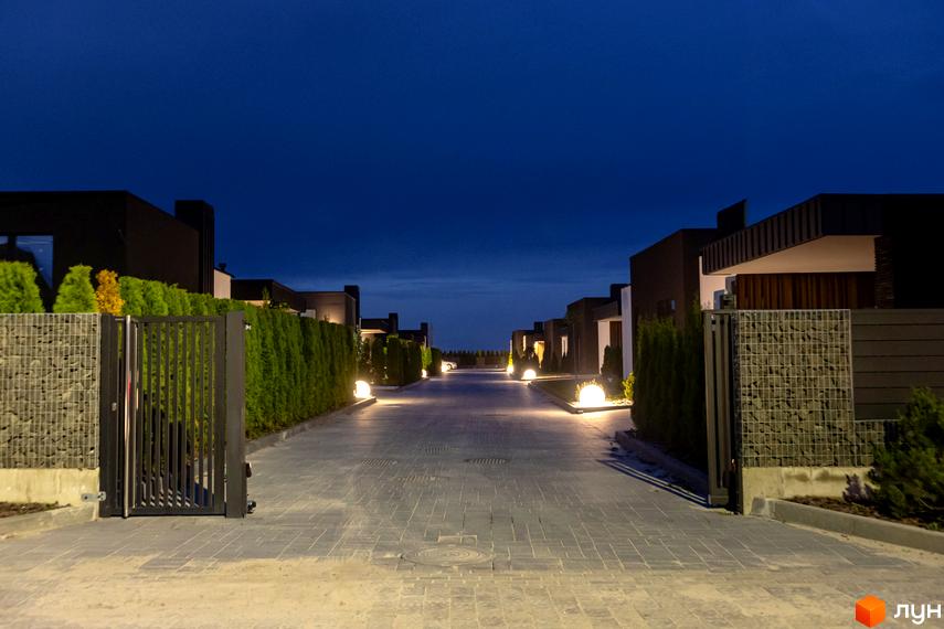 В'їзд до Містечко котеджів Terra Solis з темними будинками та брукованою дорогою ввечері. По боках зелені насадження та декоративні огорожі.