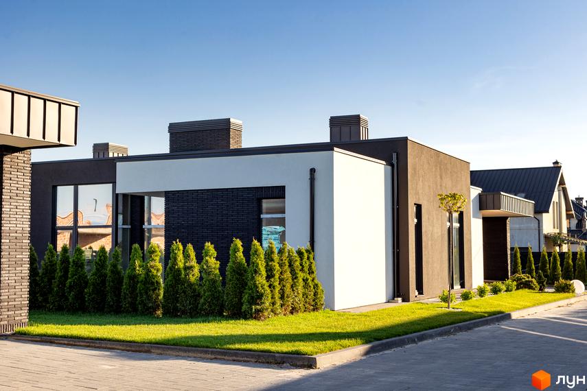 Будинок з білими та темно-сірими фасадами. Перед будинком газон з туями, бруківка та блакитне небо.