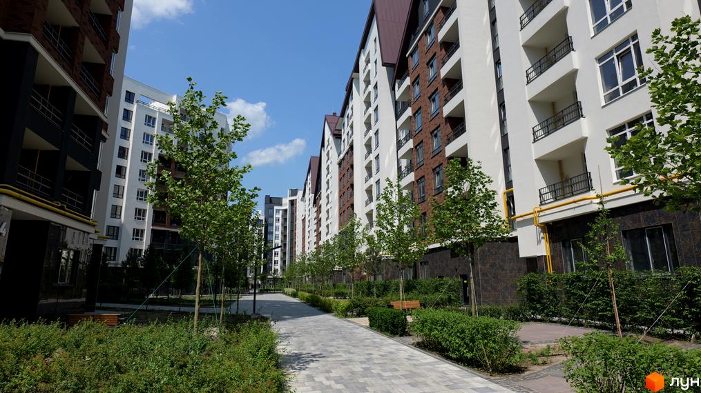 Житловий комплекс Синергія Сіті з білими та цегляними фасадами, балконами та зеленими деревами на прибудинковій території.