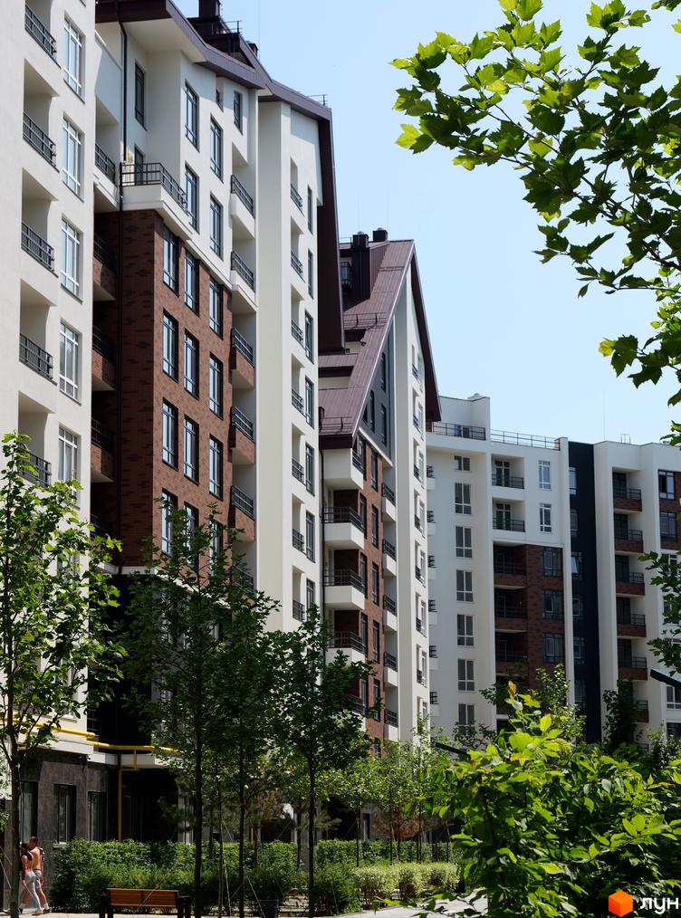 Житловий комплекс Синергія Сіті - фасади будинків з цегляними та білими елементами, балконами та зеленими деревами на прибудинковій території.