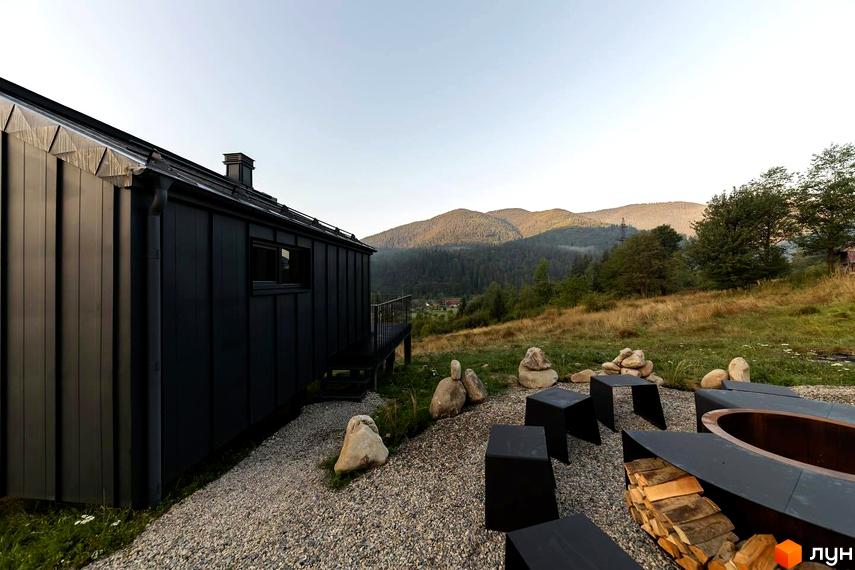 Котеджне містечко Barnhouse з чорним фасадом та терасою на тлі гірського пейзажу. На прибудинковій території розташовані місця для відпочинку з вогнищем та дерев'яними меблями.