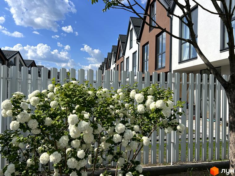 Ряд будинків Житлового комплексу Sanville Park з коричневими та білими фасадами, перед якими білий паркан та пишні кущі з білими квітами.