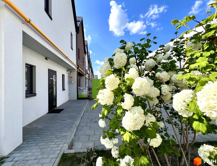 Біла будівля ЖК Sanville Park з темними вікнами та дверима, бруківкою на тротуарі та кущами з білими квітами.