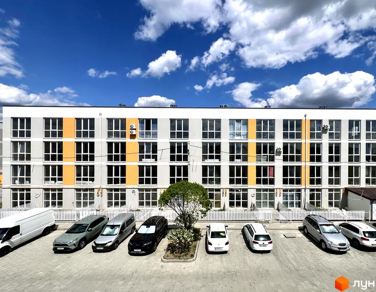 Фасад багатоповерхового будинку ЖК Sanville Park з великими вікнами та помаранчевими вставками. На передньому плані парковка з автомобілями та невелике дерево.