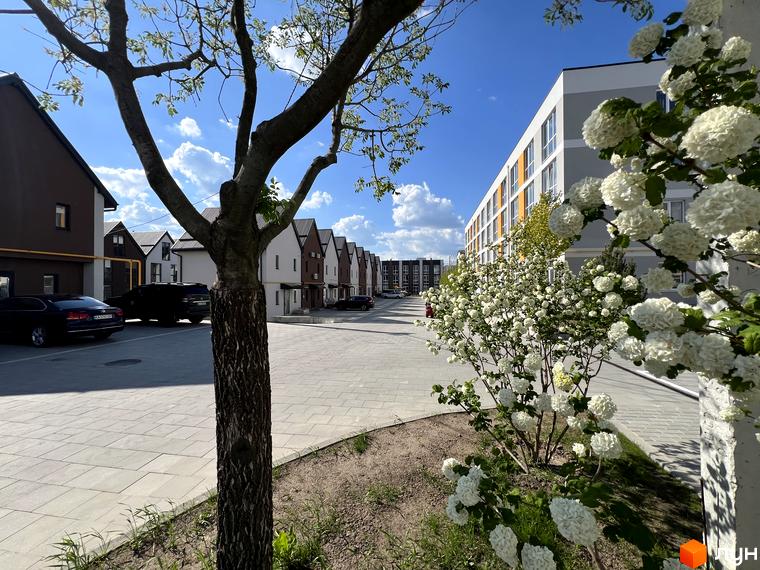 Житловий комплекс Sanville Park: ряд двоповерхових будинків та багатоповерхова будівля. Прилегла територія з квітучими кущами та вимощеною плиткою дорогою.