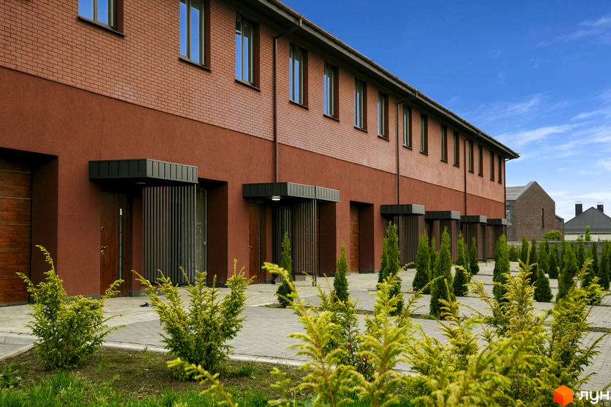 Зовнішній вигляд клубного будинку ЖК Обрій з червоної цегли та коричневої штукатурки, перед будинком висаджені туї та кущі.