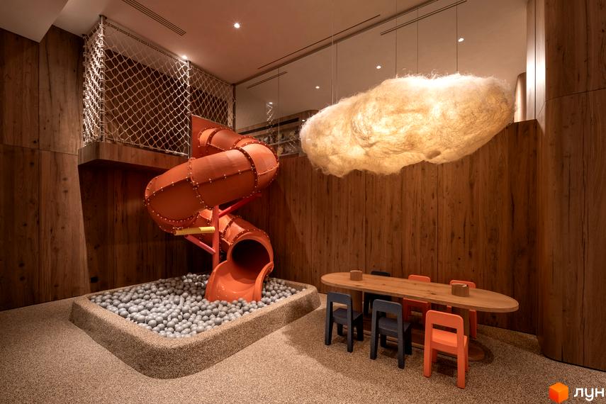 Дитяча ігрова зона в КМ Mountain Residence з гіркою та басейном з кульок, поруч стіл для дітей. Загальний вигляд інтер'єру з дерев'яними панелями та хмароподібним світильником.
