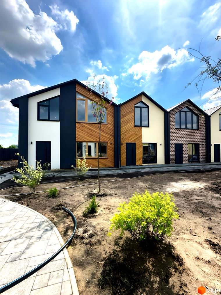 Зовнішній вигляд таунхаусів житлового комплексу PRO MISTO з панорамними вікнами та фасадами з дерева, цегли та штукатурки. На передньому плані – доглянута прибудинкова територія з рослинами.