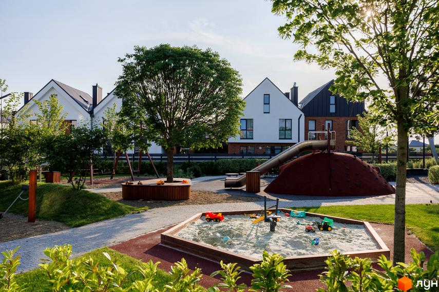 Будинки Котеджного містечка LAGOM мають білі фасади та темні дахи. На передньому плані облаштований дитячий майданчик з пісочницею та гіркою.