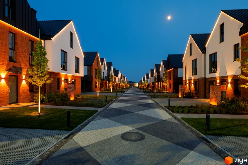 Котеджне містечко LAGOM з цегляним та білим фасадом, підсвіченим ввечері, з доглянутою прибудинковою територією та доріжкою з бруківки.