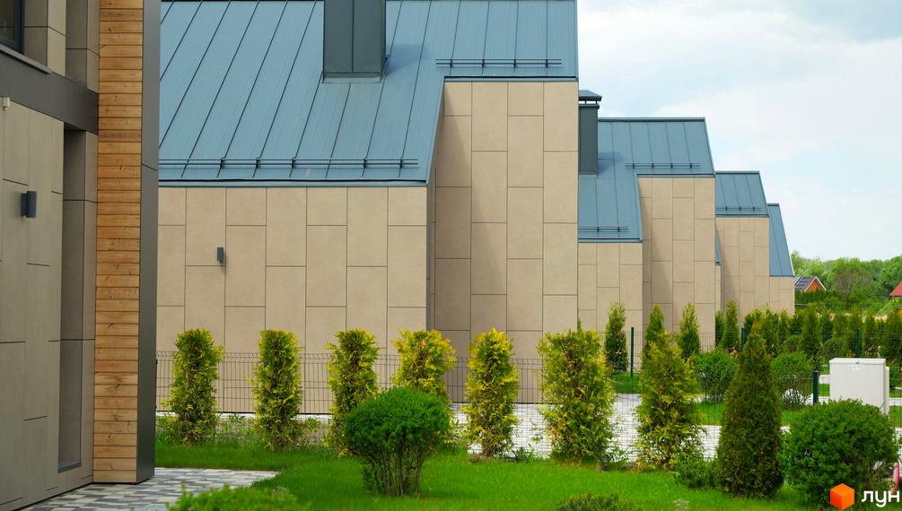 Котеджне містечко КМ Piemont eco village з фасадами з світлої плитки та металевими дахами, оточене доглянутим зеленим ландшафтом з туями та кущами.