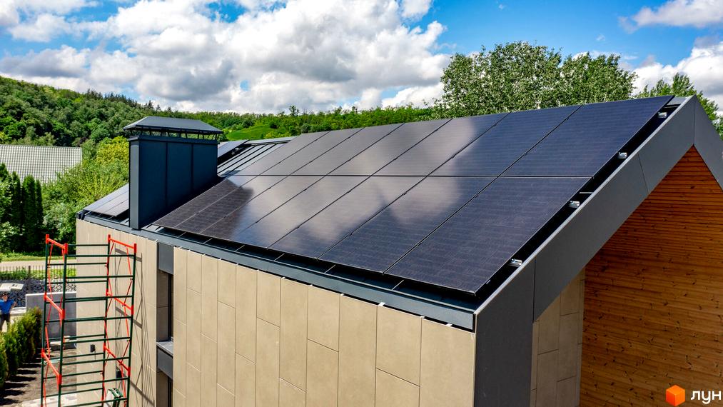 Будинок Котеджного містечка Piemont eco village з темним дахом, вкритим сонячними панелями, світлими стінами та дерев'яною обшивкою. Навколо зелені насадження.