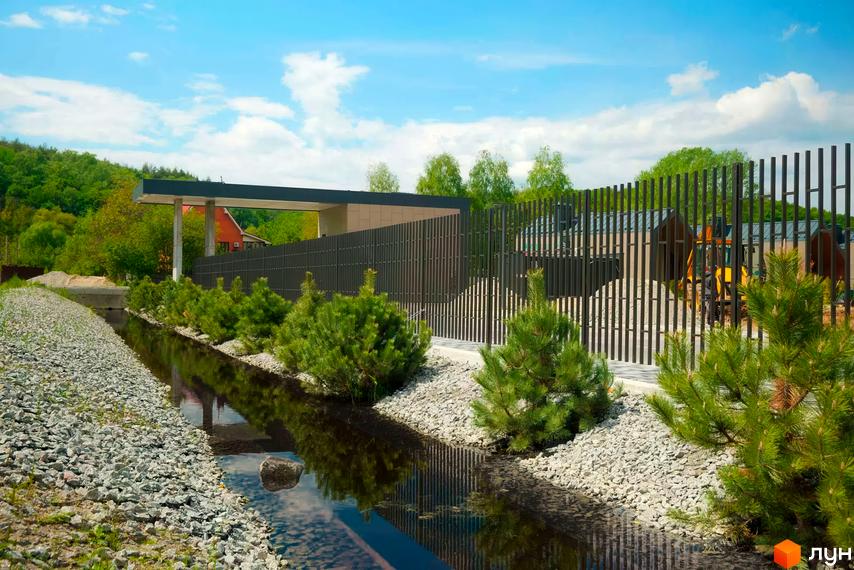 Будівлі Котеджного містечка Piemont eco village з темними дахами та навісом, за парканом, поруч з водним каналом та хвойними кущами.