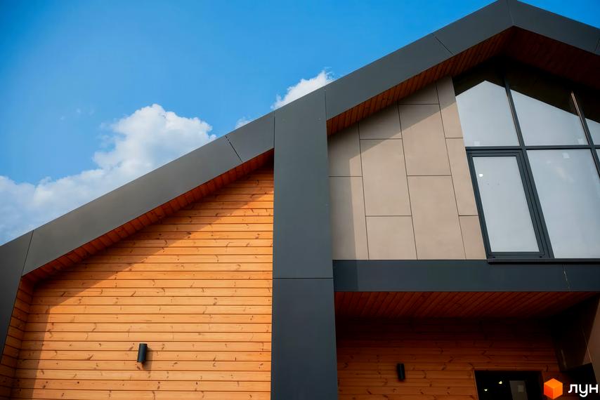 Котеджне містечко КМ Piemont eco village. Зовнішній вигляд будинку з дерев'яним фасадом та великим вікном під блакитним небом.