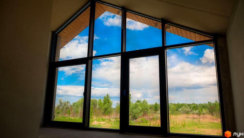 Котеджне містечко КМ Piemont eco village з панорамними вікнами, що виходять на зелену галявину та блакитне небо з хмарами.