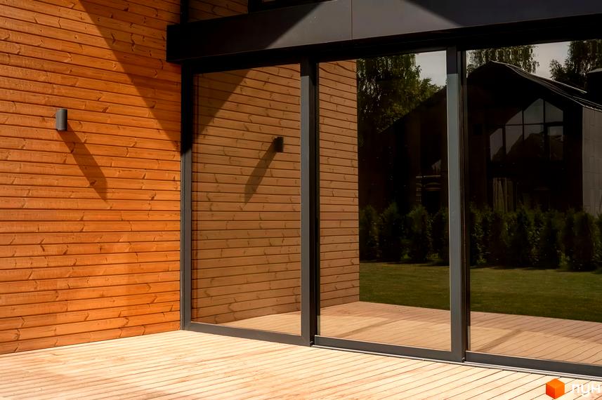 Фасад будинку Котеджного містечка Piemont eco village з дерев'яною обшивкою та великими скляними розсувними дверима. На передньому плані дерев'яна тераса, а за склом видно зелений газон.