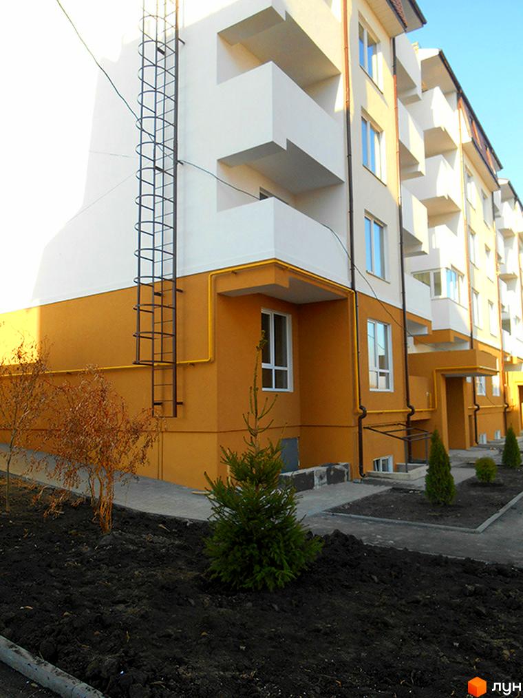 Житловий комплекс Затишне містечко. Фасад будинку з балконами та вікнами, зовнішня драбина та жовті комунікації. Прибудинкова територія з деревами та землею.