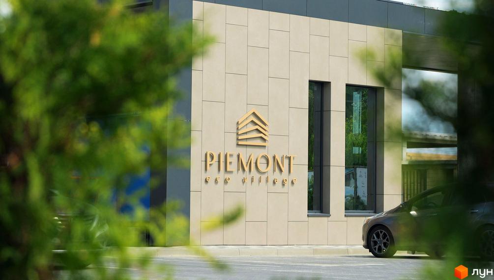 Котеджне містечко Piemont eco village з золотим логотипом та написом на фасаді. Навколо будівлі зелень та припаркований автомобіль.