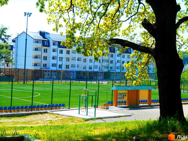 Житловий комплекс Затишне містечко з фасадом біло-жовтого кольору та блакитними дахами. Поруч розташований футбольний стадіон з трибунами та спортивний майданчик.