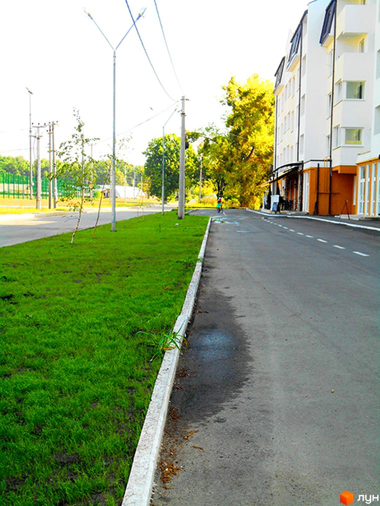 Житловий комплекс Затишне містечко: біла багатоповерхівка з жовтими елементами та доглянута прибудинкова територія з зеленою травою та дорогою.