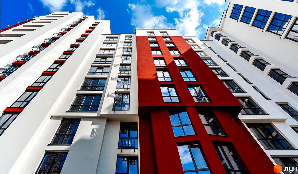 Фасад житлового комплексу Comfort House з червоними та білими елементами та великою кількістю вікон. Будинок височіє на фоні блакитного неба з хмарами.