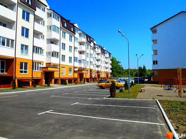 Житловий комплекс Затишне містечко з білими та жовтими фасадами, балконами та парковкою. Поруч дитячий майданчик та зелені насадження під блакитним небом.